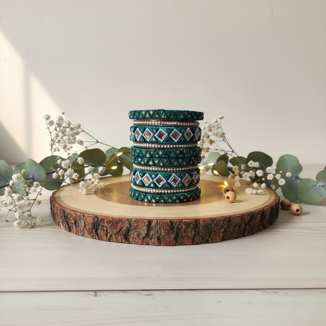 A traditional deep emerald green silk thread bangle set decorated with intricate geometric mirror work, tiny white seed beads, and gold diamond patterns, displayed stacked vertically on a rustic wooden slice surrounded by white baby's breath flowers and eucalyptus leaves.