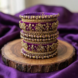 A royal purple silk thread bridal bangle set adorned with intricate gold floral Kundan work, shimmering rhinestone chain borders, and textured metallic accents, displayed stacked on a rustic wooden slice with draped purple fabric in the background.