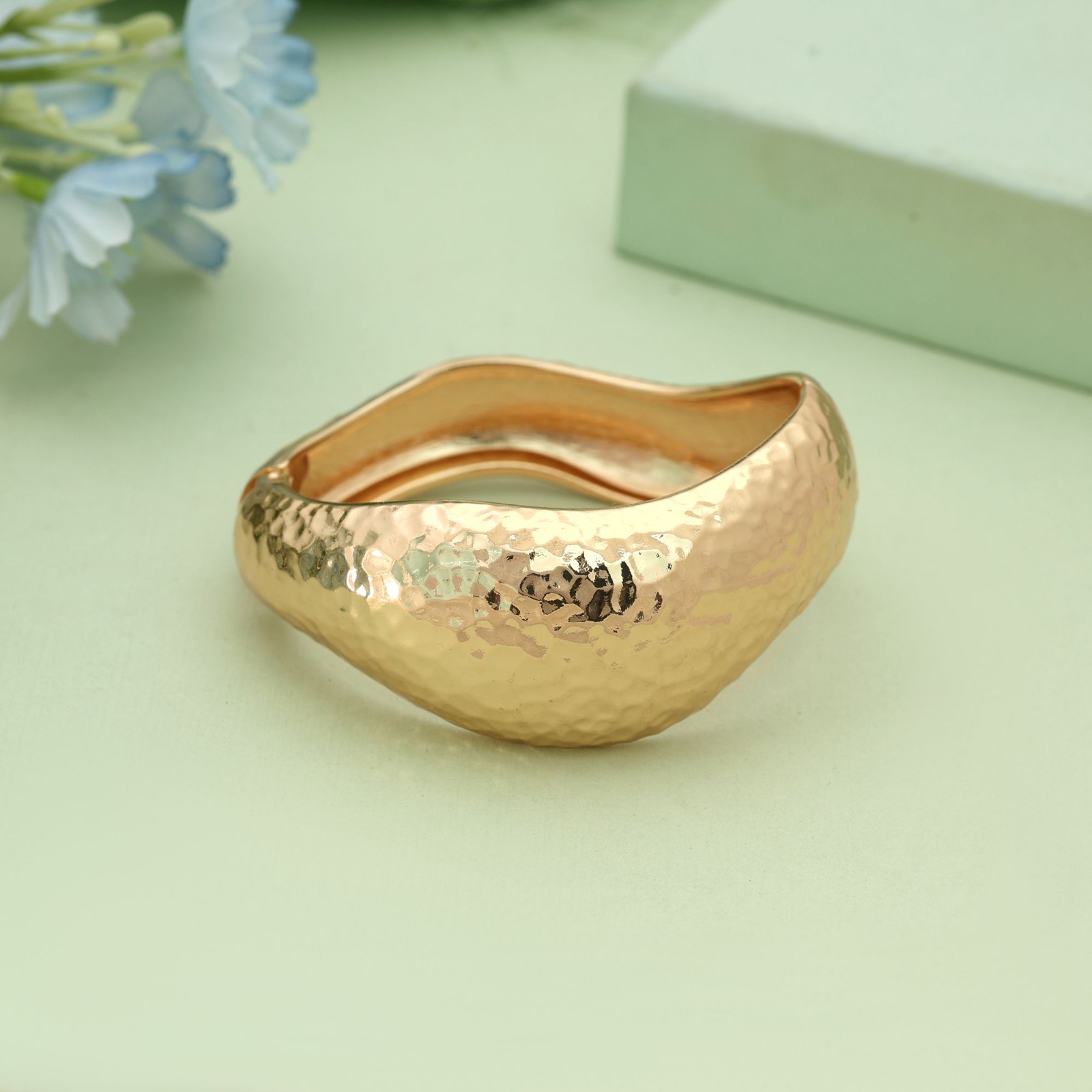 Wide gold-toned hinged cuff bracelet with a hammered texture and an organic wave shape displayed on a soft green background.