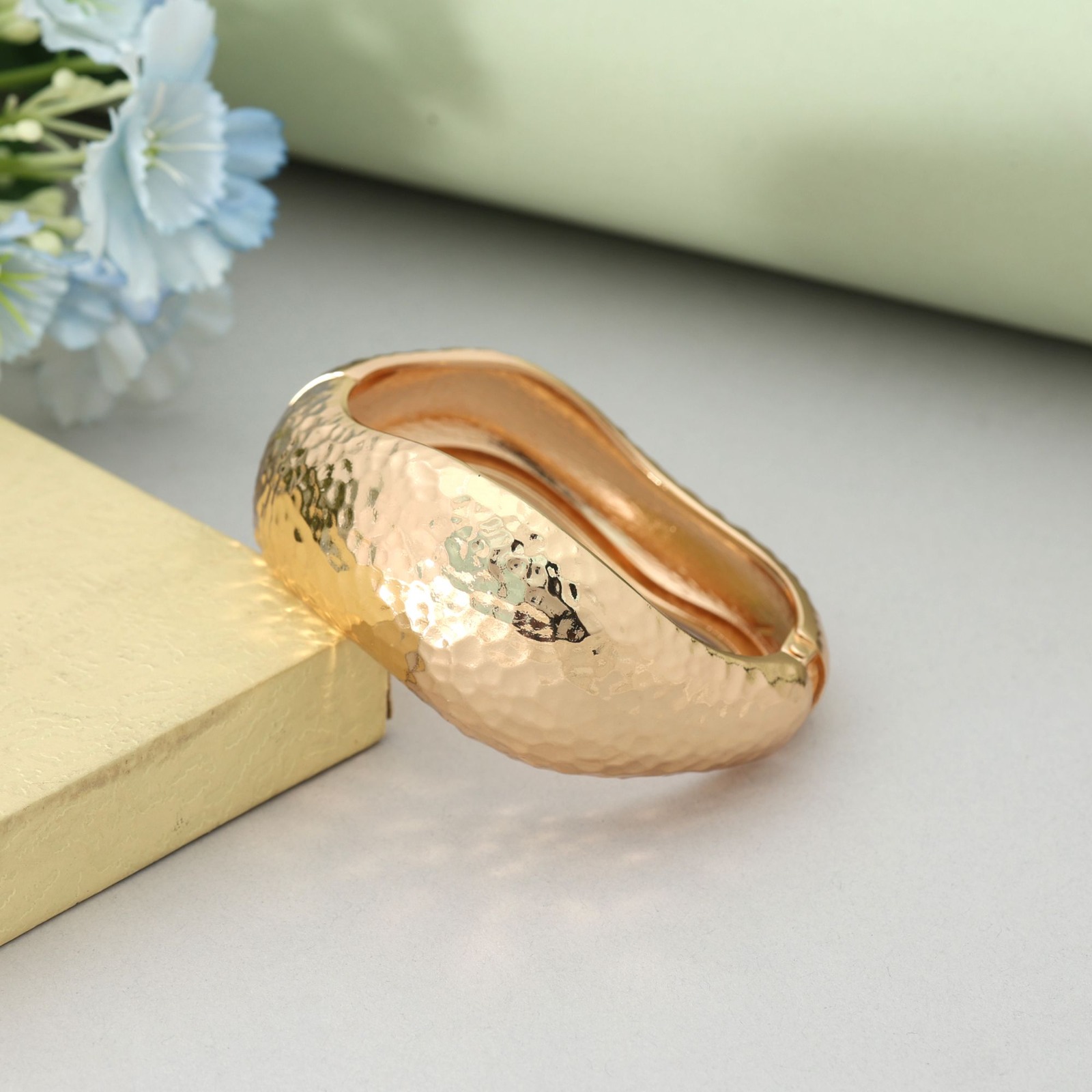 Wide gold-toned hinged cuff bracelet with a hammered texture and an organic wave shape displayed on a soft green background.