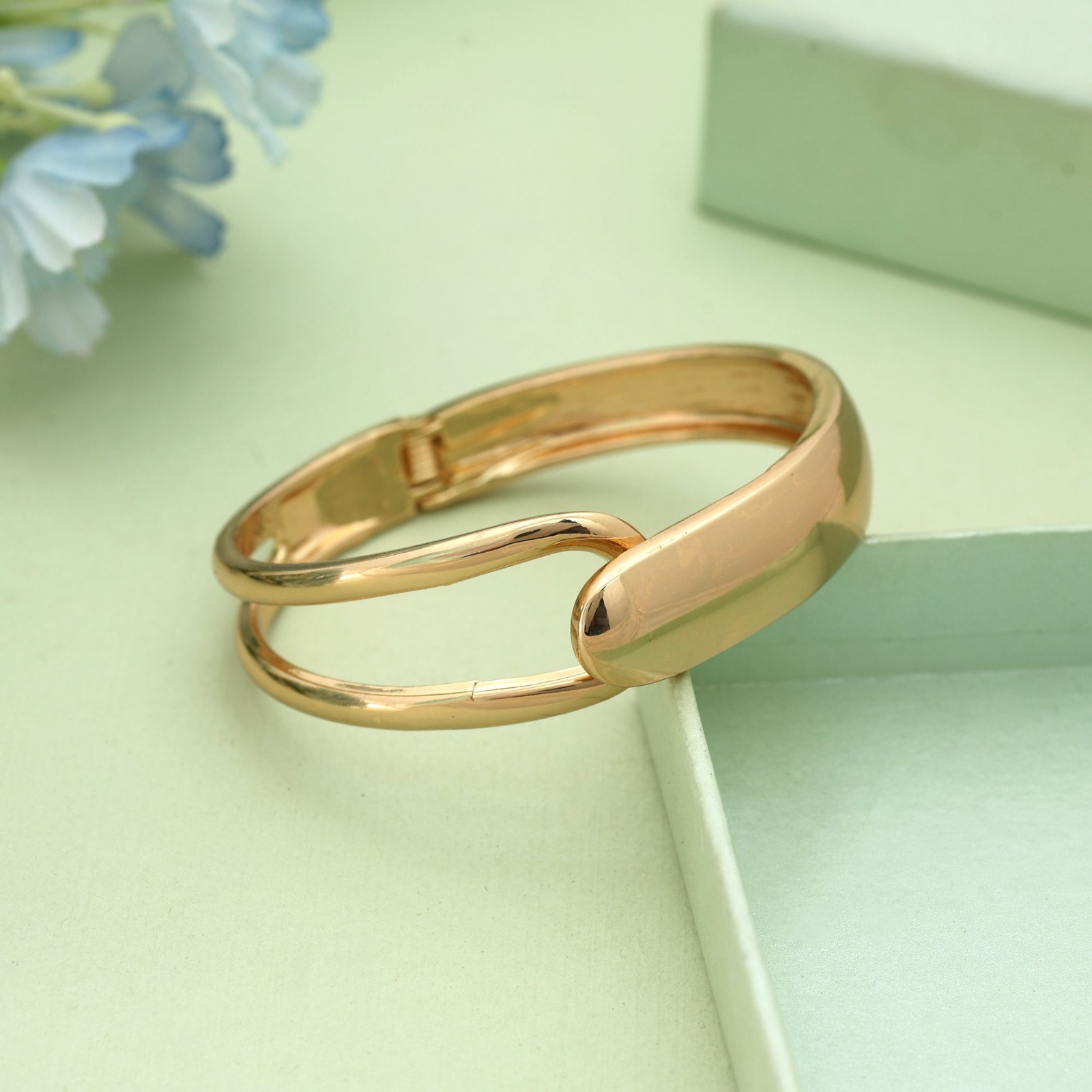 Gold-toned hinged cuff bracelet featuring an asymmetrical split-band design with a solid polished overlapping cap, resting on a soft green surface.