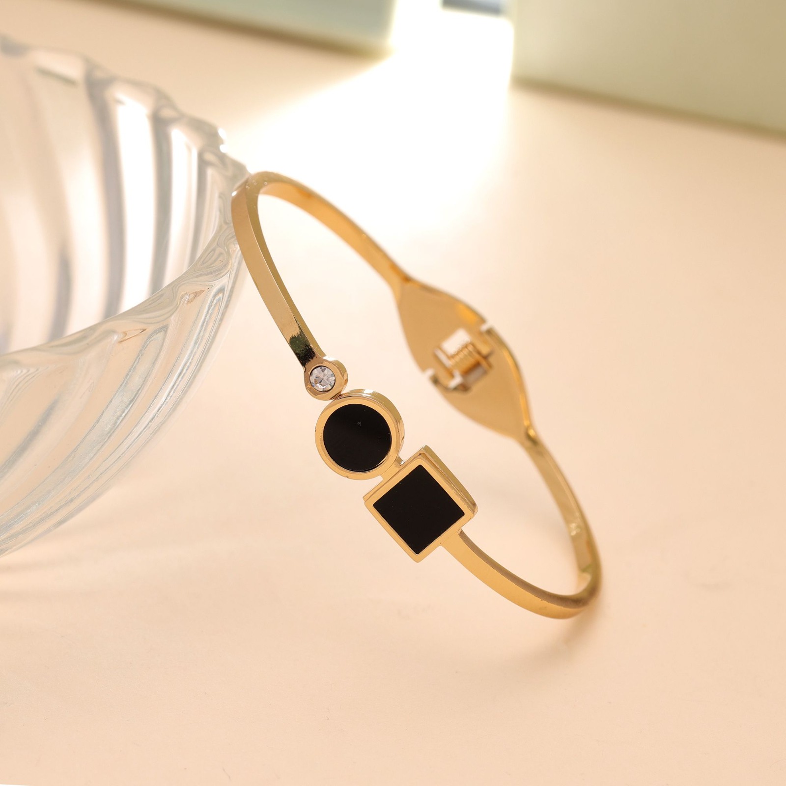Slim gold-toned hinged bangle bracelet featuring a small round crystal next to a black enamel circle and square charm, resting on a white marble block.