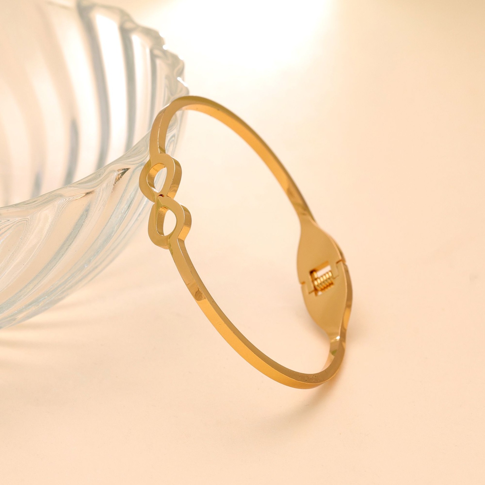 Slim gold-toned hinged bangle bracelet featuring an open infinity cutout design resting against a ribbed glass bowl on a beige surface.
