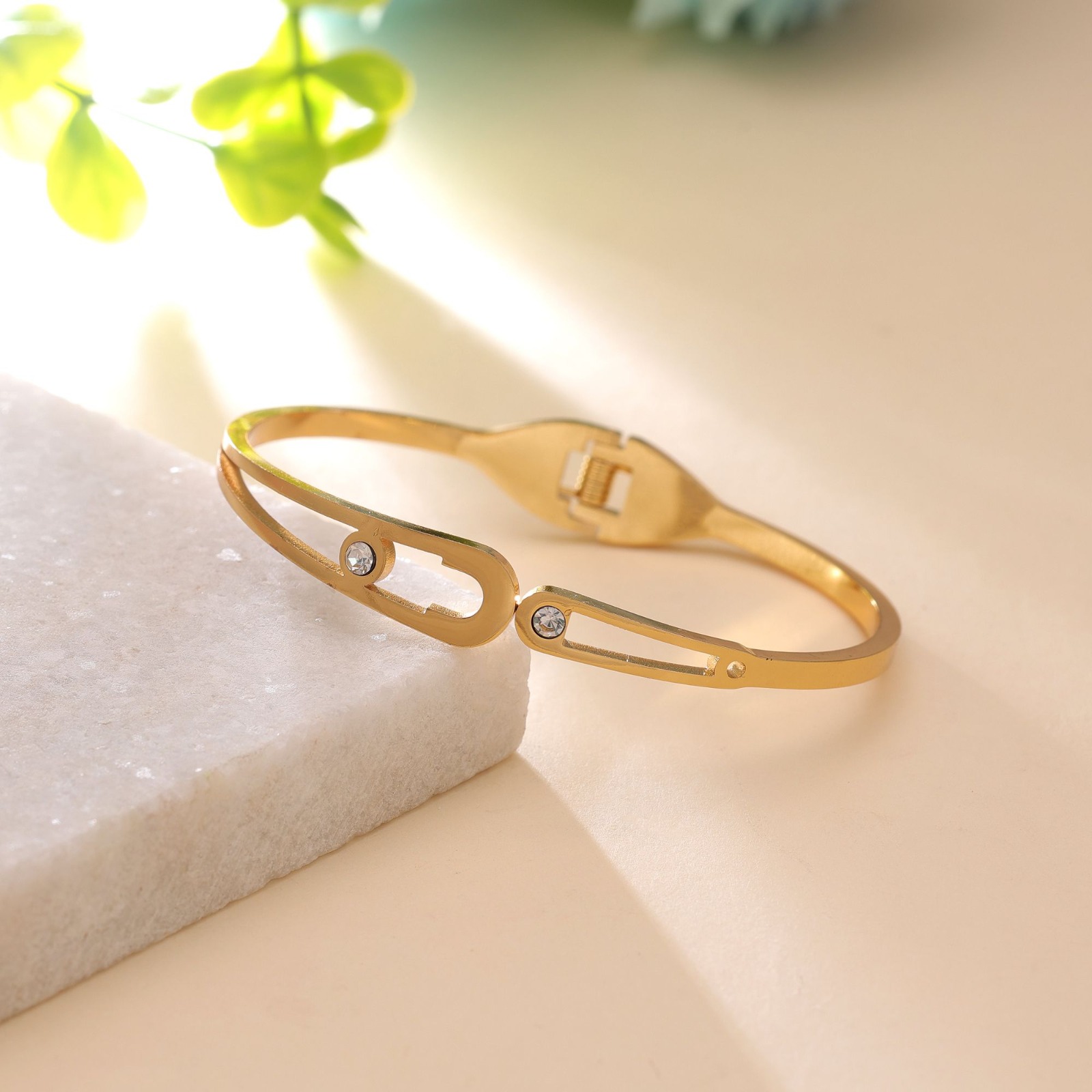 Slim gold-toned hinged bangle bracelet featuring dual oval cutout motifs with sparkling bezel-set crystals, resting on a white marble slab.