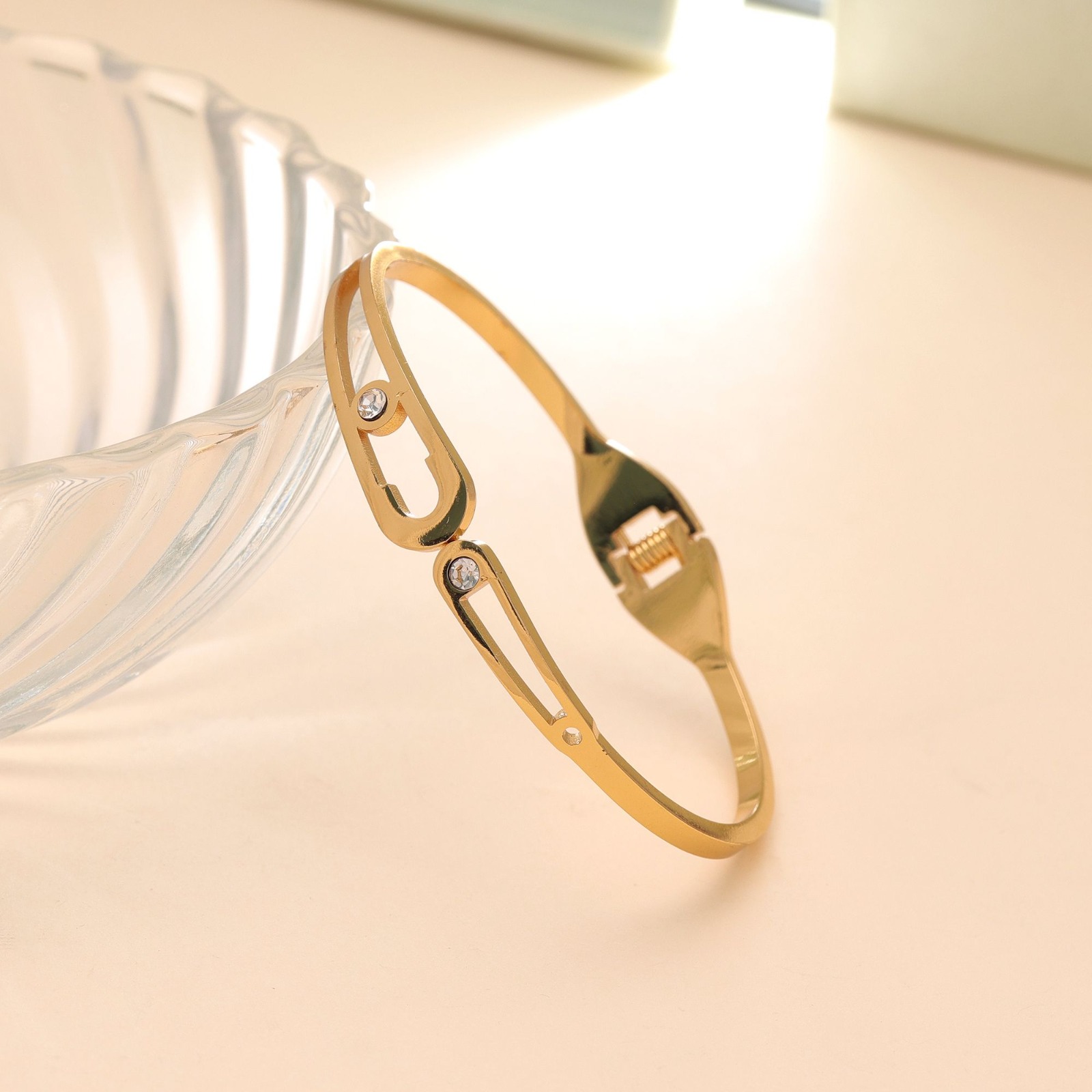Slim gold-toned hinged bangle bracelet featuring dual oval cutout motifs with sparkling bezel-set crystals, resting on a white marble slab.