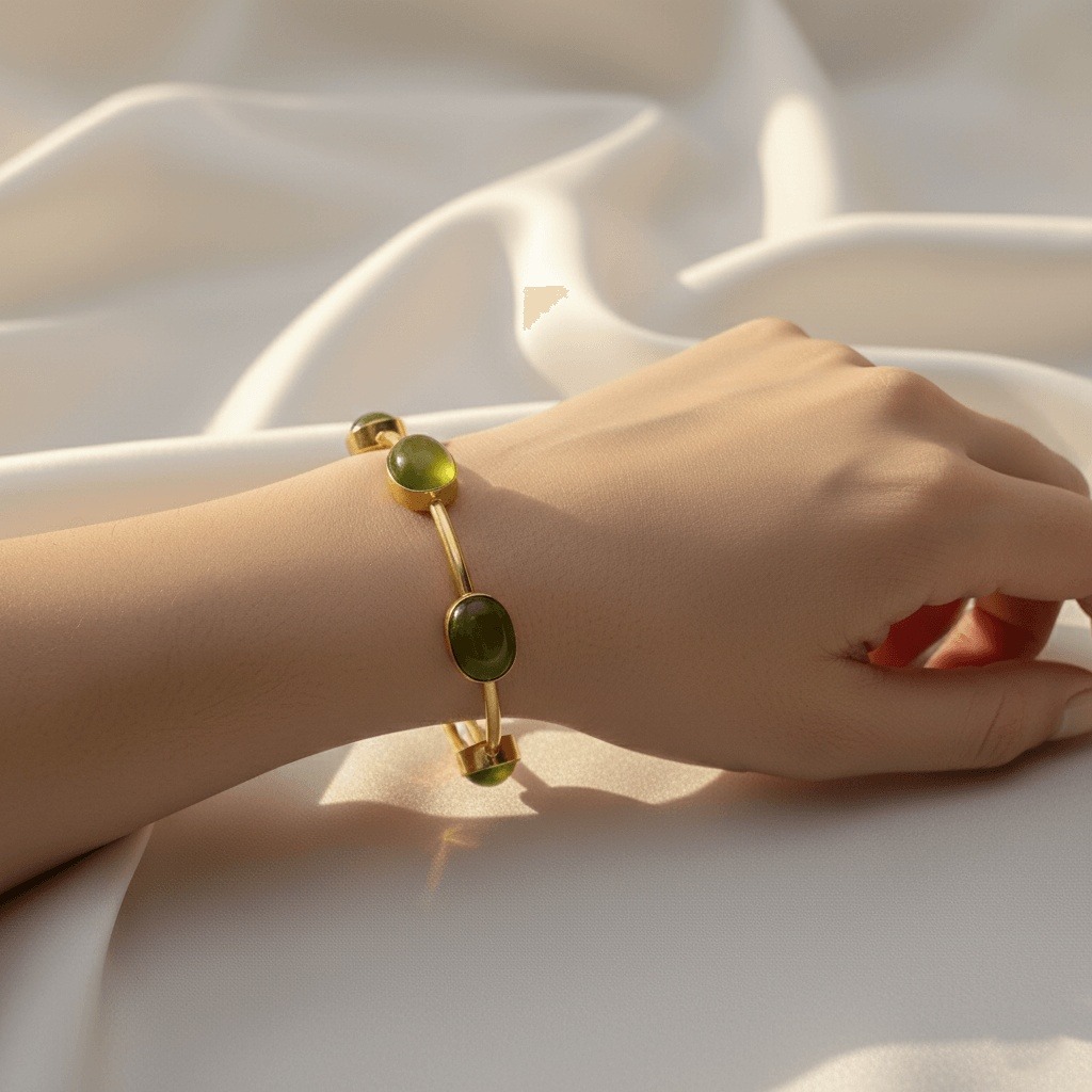 Gold-toned open cuff bangle featuring four oval olive-green monalisa stones bezel-set along a slender wire band, resting on a textured white fabric background next to a white orchid flower.